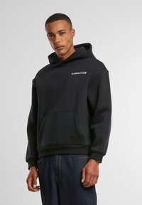 Zwarte hoodie van katoen met een voorste kangoeroezak, ribgebreide manchetten en de tekst "KARMA CLUB" in het wit op de linkerbovenkant van de borst.