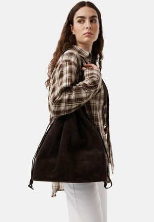 Donkerbruine suède handtas met een slouchy ontwerp, trekkoordsluiting en contrasterende lichte stiksels. Draagt over de schouder bij een casual outfit.