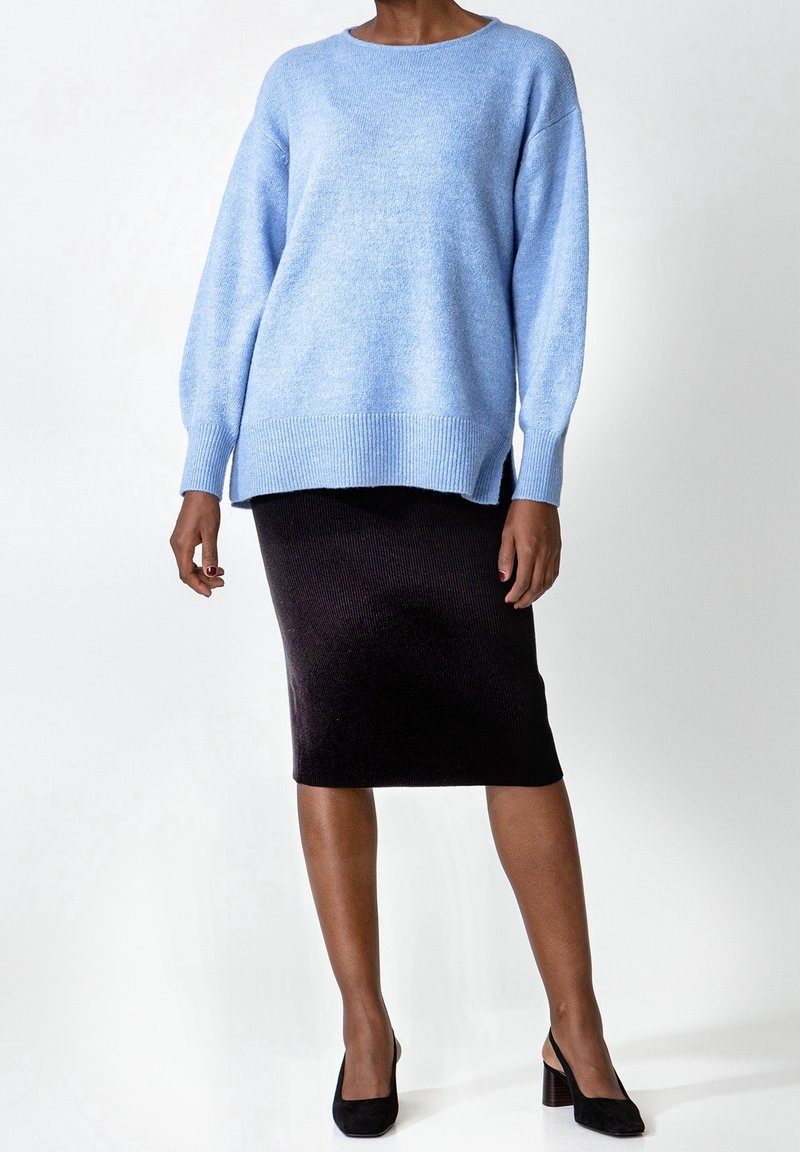 Personne portant un pull bleu clair à manches longues, une jupe crayon noire jusqu'aux genoux, et des chaussures à talons noires sur fond blanc.