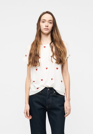 Junge Frau mit langen braunen Haaren, die ein weißes T-Shirt mit kleinen roten Herzmustern und eine dunkelblaue Jeans trägt, vor einem schlichten Hintergrund stehend.