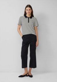 Gestreept poloshirt met een zwarte kraag, gecombineerd met zwarte cropped broeken. Model staat met handen in de zakken, en draagt zwarte slingback flats.