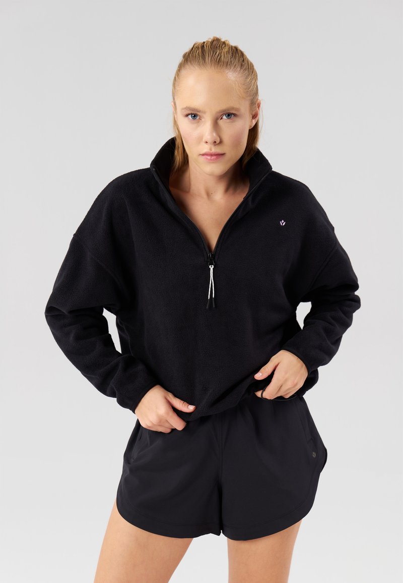 Černý fleecový pulovr se zipem u krku, s malým logem na hrudi. Kombinován s černými šortkami z lehké tkaniny.