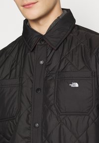 Gros plan d'une personne portant une veste matelassée noire avec fermeture par boutons et poches poitrine, avec le logo The North Face sur la poche droite.