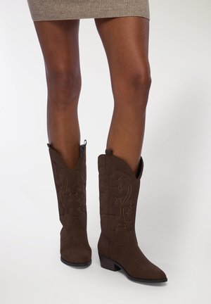 Stivali da cowboy in suede marrone con punta appuntita, tacco basso e impilato, e cuciture decorative con un motivo floreale lungo il gambale.