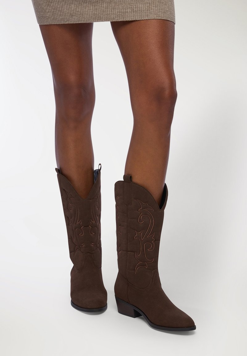 Stivali da cowboy in suede marrone con punta appuntita, tacco basso e impilato, e cuciture decorative con un motivo floreale lungo il gambale.