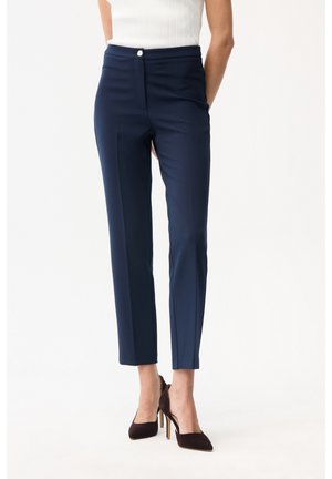 Kobieta ubrana w dopasowane, granatowe spodnie kończące się przy kostkach oraz czarne buty na wysokim obcasie, stojąca z rękami za plecami.