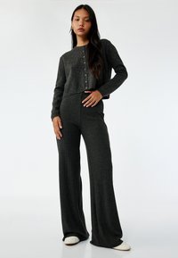 Grå strikket cropped cardigan med knaplukning, parret med brede grå ribbede bukser. Glat tekstur; model står oprejst foran en hvid baggrund.