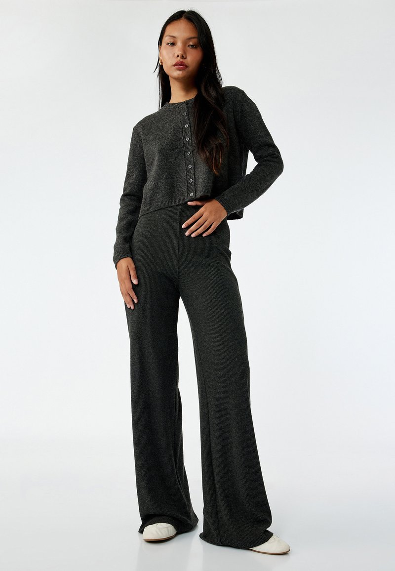 Grå strikket cropped cardigan med knaplukning, parret med brede grå ribbede bukser. Glat tekstur; model står oprejst foran en hvid baggrund.