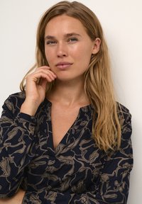 Marineblauwe shirt met een vloeiende, beige abstract patroon, voorzien van een diepe V-hals en lange mouwen. De stof lijkt zacht en lichtgewicht te zijn.
