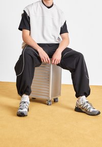 Harmaa hihaton paita mustan paidan päällä, yhdistetty mustiin housuihin, joissa valkoiset reunukset. Harmaat ja mustat lenkkitossut. Istuu hopeanvärisell ä pyörällisellä esineellä.