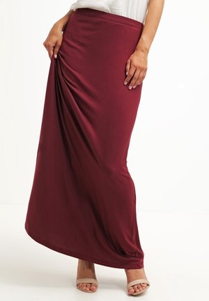 Femme portant une longue jupe bordeaux tenant le tissu sur le côté, assortie à des sandales beiges à bouts ouverts et un haut blanc, sur un fond blanc.