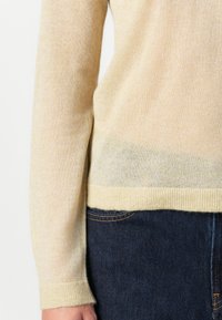 Beige langermet genser med ribbestrikket mansjett og kant, båret med mørkeblå dongeribukser, som viser arm- og hoftområdet.