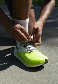 Jasně zelená sportovní bota s hladkým povrchem, černými tkaničkami a bílou podrážkou. Ruce zavazují tkaničky na dlážděném povrchu.