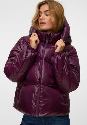 Femme ajustant la capuche d'une doudoune violette brillante, portant un jean bleu, sur un fond clair uni.