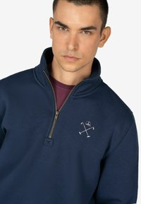 Sudadera azul marino con cremallera fabricada en un tejido suave, que presenta un cuello con cremallera de cuarto, bordado de logo y sutil herraje de cremallera marrón.