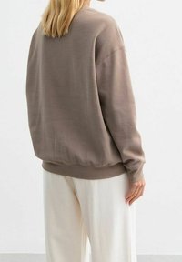 Sweatshirt in cotone marrone con vestibilità rilassata, polsini e orlo a coste e spalle scese. Abbinato a pantaloni bianchi a gamba larga.