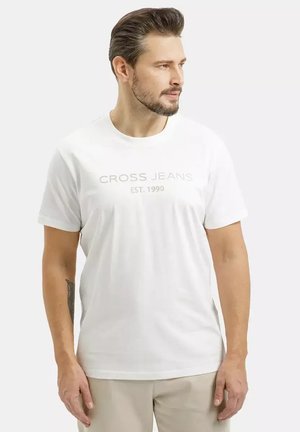 Mężczyzna z brodą w białej koszulce "CROSS JEANS EST. 1990" i beżowych spodniach, patrzy w prawo na jednolitym białym tle.