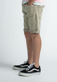 Beige denim shorts met opgerolde boorden, voorzien van verticale zakken en een achterzak met label, gecombineerd met zwarte Vans schoenen met witte accenten.