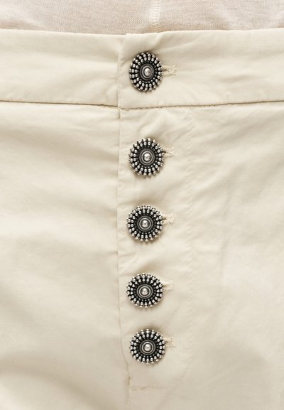 Μπεζ ύφασμα με πέντε στρογγυλά, διακοσμητικά ασημένια κουμπιά ραμμένα κάθετα, καθένα με κεντρικό άνισο θόλο και περιμετρικές λεπτομέρειες με χάντρες.