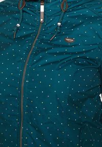 Teal waterproof jacket featuring multicolored polka dots, zippered front, and drawstring hood with brown accents and logo patch.