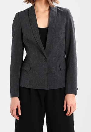 Donna che indossa un blazer grigio scuro a un bottone sopra una maglietta nera e pantaloni neri su uno sfondo chiaro uniforme.
