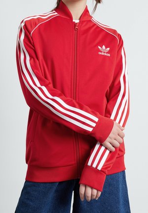 Person iført en rød Adidas lynlåsjakke med hvide striber på ærmerne, logo på brystet og blå denimbukser, med armene krydset ved taljen.