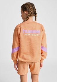 Jasnopomarańczowa bluza z fioletowym paskiem, z logo "hummel" i "SPORTSWEAR" z tyłu, w zestawie z pasującymi szortami.