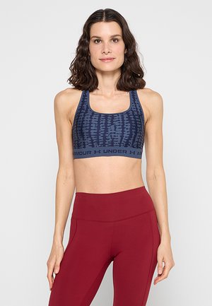 Vrouw draagt een blauw patroon Under Armour sportbeha en rood hoog getailleerde leggings, staand tegen een effen lichte achtergrond.