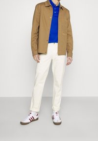 Giacca marrone con bottoni, polo blu sotto, pantaloni bianchi a gamba dritta e sneakers bianche a strisce rosse. Design semplice con cuciture visibili.