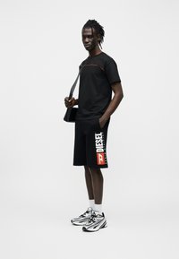 Μαύρο μπλουζάκι με κόκκινη λεπτομέρεια, μαύρο σορτς με λευκό και πορτοκαλί λογότυπο "DIESEL", και ασημί αθλητικά παπούτσια με μαύρες λεπτομέρειες.