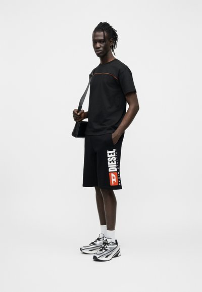 T-shirt noir avec accent rouge, short noir arborant le logo "DIESEL" en blanc et orange, et baskets argentées avec accents noirs.
