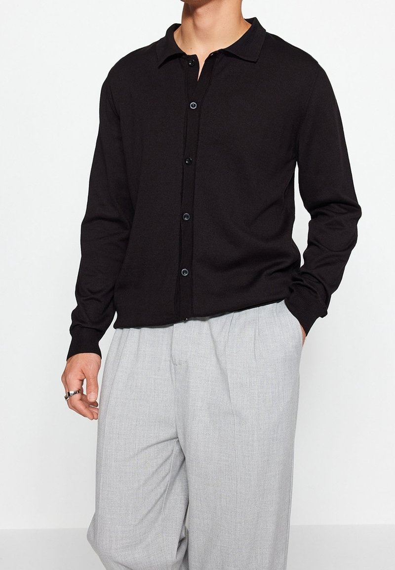 Svart cardigan med knäppning framtill, krage, långa ärmar och ribbade manschetter. Parat med löst sittande ljusgrå byxor.