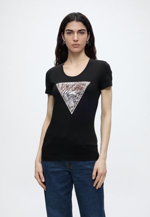 Femme aux longs cheveux foncés portant un t-shirt noir ajusté avec un grand logo triangulaire à motifs et un jean bleu, debout devant un fond uni.
