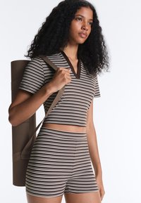 Jeune femme portant un tapis de yoga dans le dos, vêtue d’un crop top et d’un short rayés marron et blanc assortis, regardant devant elle.