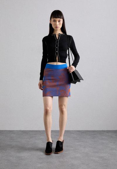 Cardigan negru cu mâneci lungi și detalii albe, asociat cu o fustă mini cu un model albastru și portocaliu. Pantofi negri cu un design unghiular. Geantă neagră.