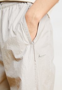 Svetlo sivi Nike trenirka s elastičnim pasom, stranskimi žepi in majhnim srebrnim logotipom na stegnu. Teksturni, lahki material.