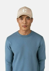 Lyseblå genser med rund hals og ribbet tekstur, kombinert med en beige baseballcap med et hvitt brodert "C"-logo.