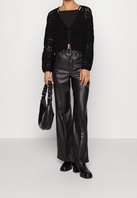 Černý háčkovaný cardigan s detailem na zavazování, v kombinaci s černými kalhotami z umělé kůže s vysokým pasem a zmačkanou černou kabelkou. Tlusté kozačky na podpatku.