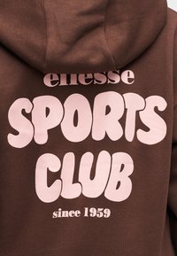 Sweat à capuche marron avec texte rose indiquant "ellesse SPORTS CLUB depuis 1959" au dos, montrant la texture du tissu et les détails de la capuche.