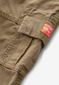 Tan cargo fabric with visible stitching and a prominent red and orange "Superdry" branded patch. Features a pocket and textured surface.