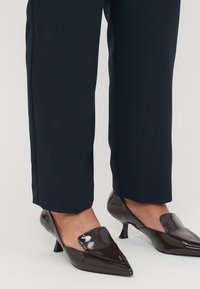 Brown patent leather mules with a pointed toe and low heel, featuring a smooth texture and a slip-on design, worn with navy trousers.