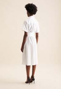 Weißes Leinenhemdkleid mit kurzen Ärmeln, tailliertem Schnitt und fließendem Rock. Schwarze Ankle-Strap-Absätze runden das Outfit ab.