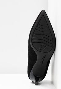 Scarpa nera in suede con tacco alto e punta affusolata, dotata di una suola in gomma decorata, con la taglia 37 visibile sul fondo.