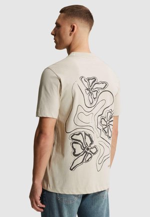 Homme portant un t-shirt beige avec un motif floral abstrait noir en ligne dans le dos, associé à un jean bleu, tourné vers la gauche sur un fond uni.