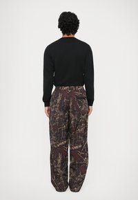 Zwarte lange mouwen shirt gecombineerd met losse bruine camouflagebroek met een gestructureerd patroon. De broek heeft een achterzak.