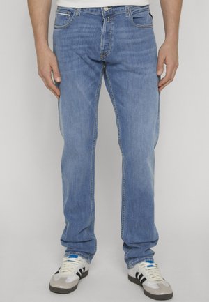 Mann trägt hellblaue Straight-Fit-Jeans und weiße Sneakers mit schwarzen Streifen, steht vor einem schlichten Hintergrund.