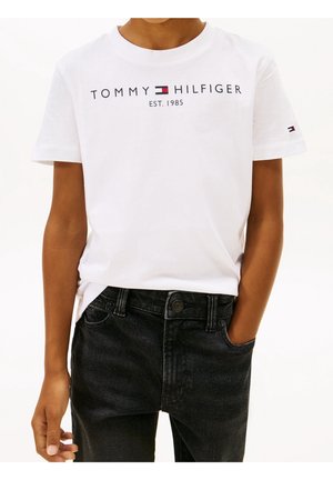Person iført en hvid Tommy Hilfiger t-shirt med logo og teksten "EST. 1985", kombineret med sorte denimjeans, den ene hånd i lommen.