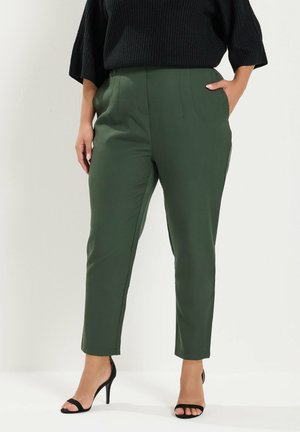 Mujer vestida con pantalones de corte ajustado de cintura alta en verde oscuro, top de punto negro y sandalias negras de tacón alto con dedos abiertos, de pie con una mano en el bolsillo.