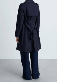 Osoba ve tmavě modrém trenchovém kabátu s páskem vzadu a širokých tmavých džínách, stojící na šedé podlaze u bílé zdi.