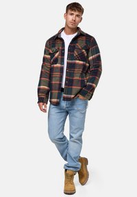 Camisa de franela a cuadros en verde oscuro, azul marino y óxido con cierre de botones, dos bolsillos en el pecho, jeans de mezclilla y botas marrones con cordones.